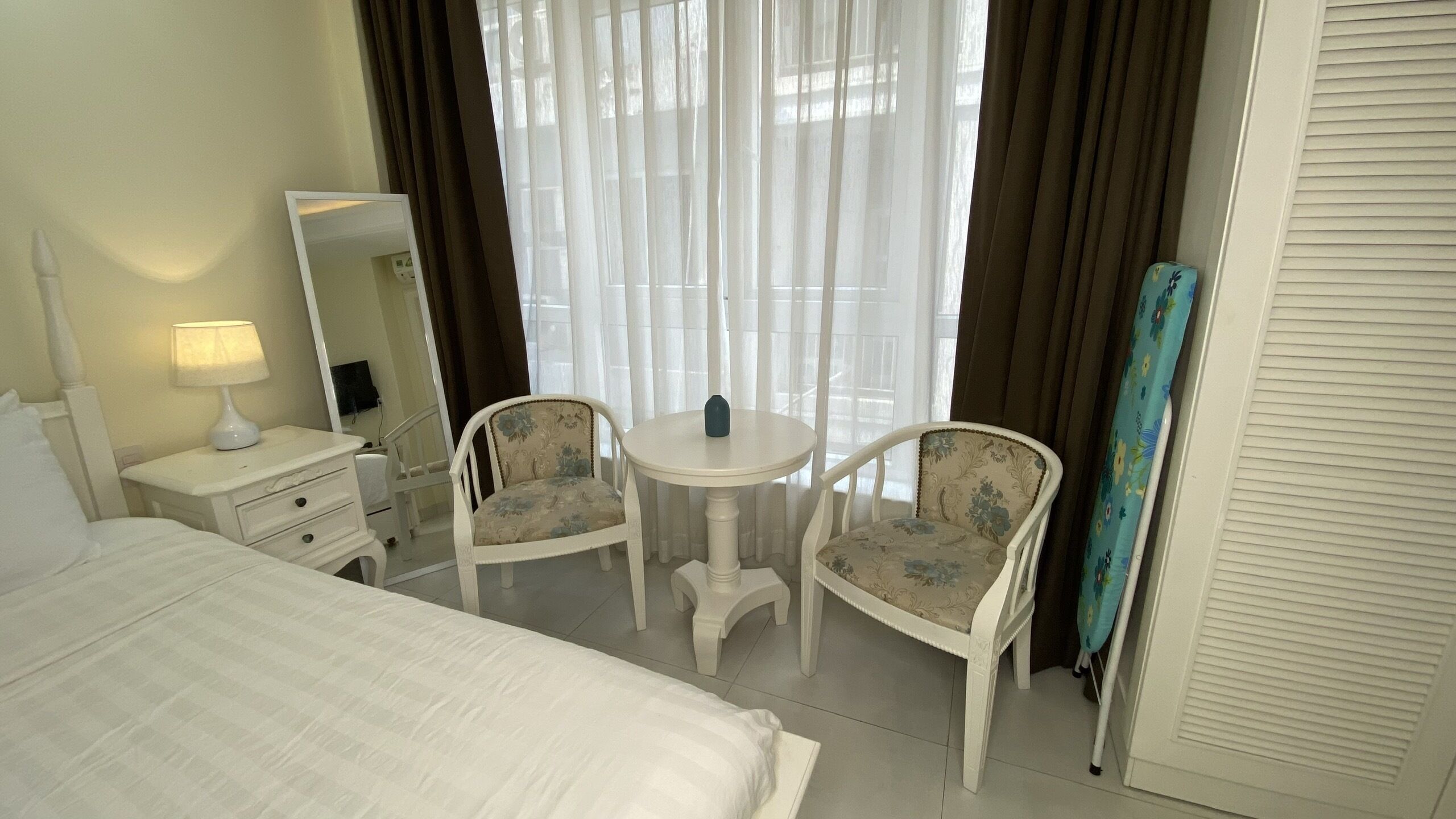Une chambre d’hôtel avec un lit, un bureau avec une lampe, deux chaises, une petite table ronde, un miroir et une fenêtre avec des rideaux.