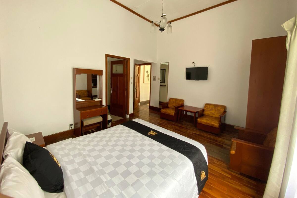 A hotel room with a large bed, a wooden dresser, a desk, two armchairs, a television, and a ceiling fan.