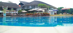 A swimming pool with a poolside area, umbrellas, and a building with a curved roof in the background. - Concordia Hotel (Bandung)