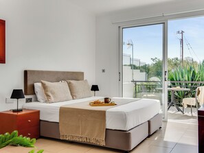 A modern bedroom with a large bed, a nightstand, a lamp, and a balcony with a view of greenery and a fence.