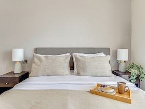 Una cama bien hecha con dos almohadas, una mesita de noche con una lámpara, y una bandeja con un tazón de cereal y una taza.