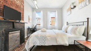 Un dormitorio moderno con pared de ladrillo, chimenea, cama con ropa de cama blanca, escritorio con silla y televisor montado en la pared.