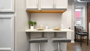Una cocina con armarios blancos, encimera blanca, dos taburetes de bar, una planta en maceta y una pared de ladrillo al fondo.