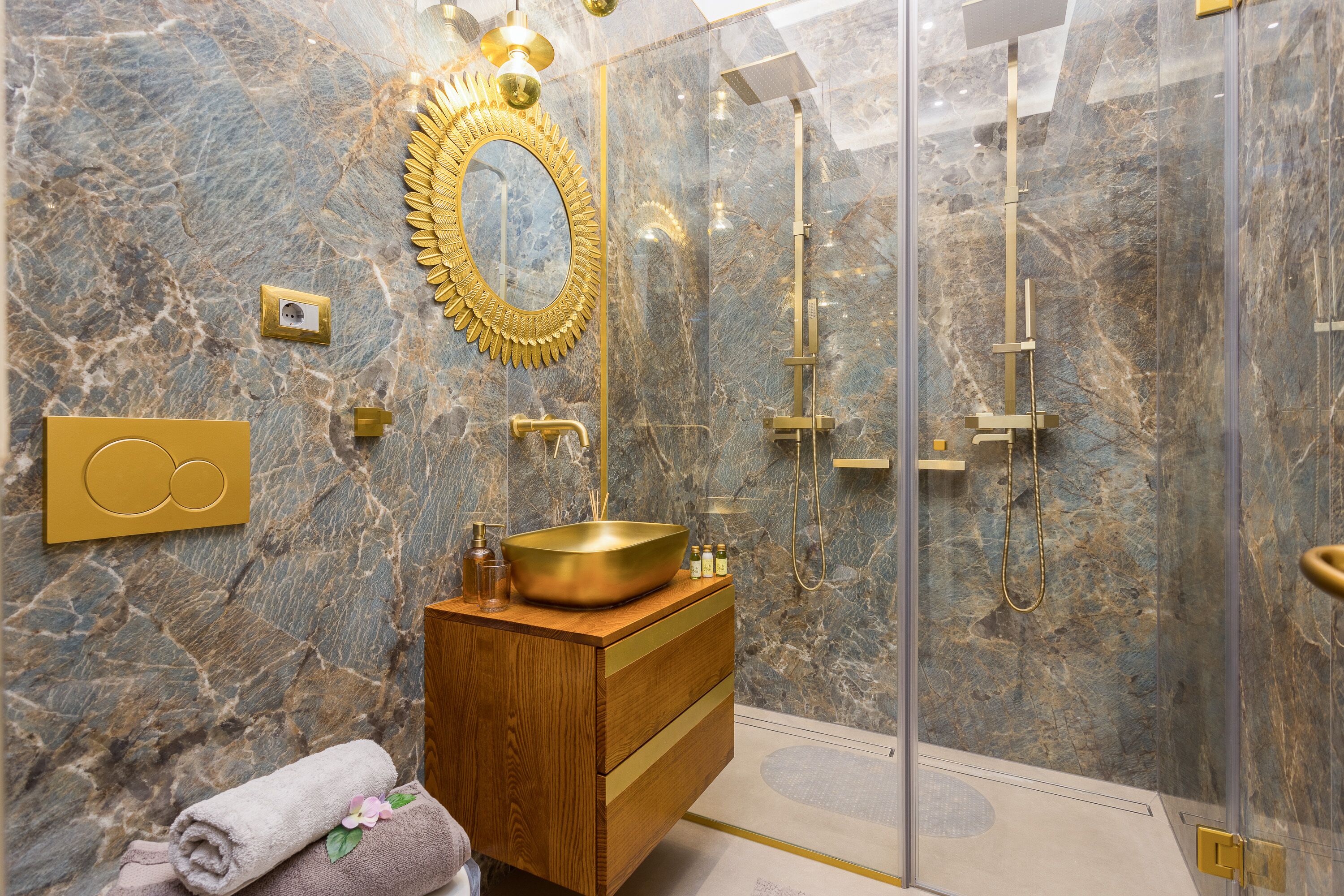Une salle de bain luxueuse avec un mur en marbre, un lavabo doré et une douche séparée par des parois en verre.