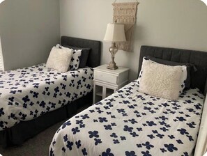 Twee eenpersoonsbedden met bloemenbedrukking, een nachtkastje met een lamp en een wanddecoratie van macramé.