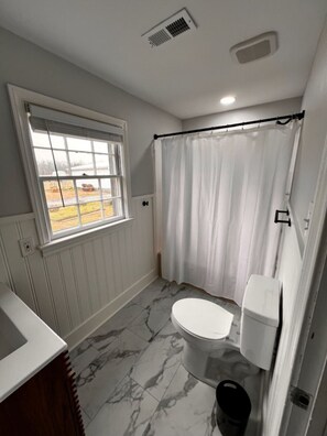 A bathroom with a toilet, a window, a shower curtain, and a marble floor. - The Cracker Jack House: A Military Salute & Arcade! (Surry)