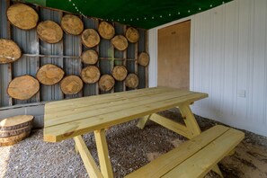 A wooden picnic table with benches outside a building with a green awning and a wall decorated with circular wooden logs. - The Cracker Jack House: A Military Salute & Arcade! (Surry)
