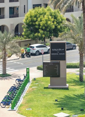 A sign for 'The Greens Sook' with a list of shop listings, a bike parking area, and a white car parked on the street. - Vogue | The Greens near Emirates Mall | Ski Dubai (Dubai)