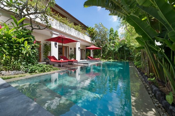 A swimming pool with lounge chairs and umbrellas, surrounded by lush greenery and a white building with balconies.
