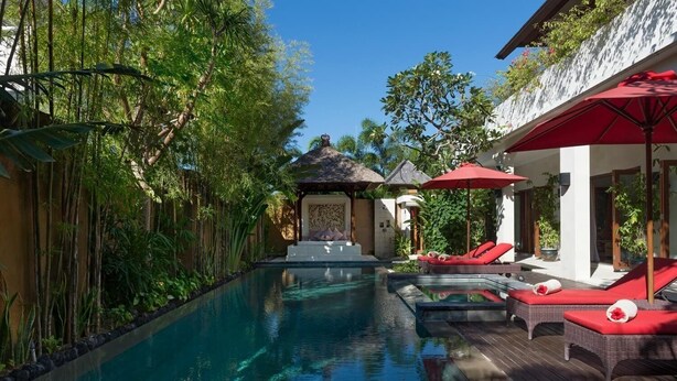 A pool area with lounge chairs and umbrellas, surrounded by lush greenery and a traditional thatched-roof structure. - Villa Kalimaya (Seminyak)
