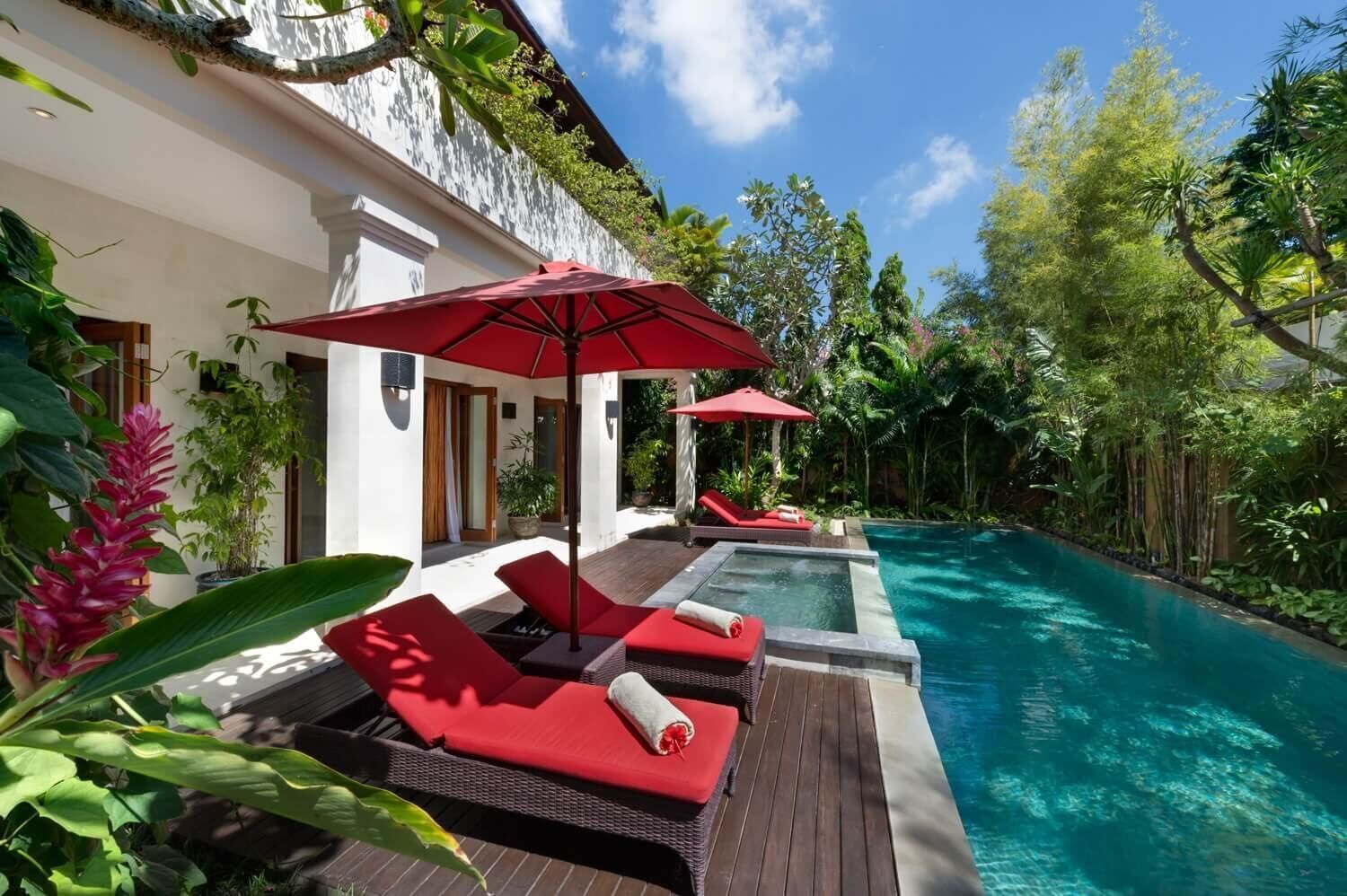Un espace aménagé au bord de la piscine, avec des chaises longues rouges et des parasols, entouré d’une végétation luxuriante et surplombant une piscine à l’eau d’un bleu limpide.