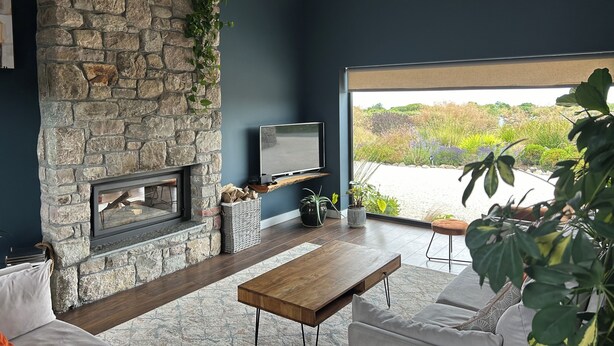 Ein modernes Wohnzimmer mit Kamin aus Stein, einem Flachbildfernseher, einem Holztisch und einem großen Fenster mit Blick auf einen Garten.