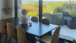 Ein moderner Essbereich mit einem großen Tisch und Stühlen, einer Vase mit Pflanze sowie Blick auf Grünflächen und das Meer durch große Fenster.