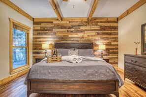 Een slaapkamer met een houten wand van boomstammen, een bed met een grijze deken, twee bedlampen en een houten dressoir.
