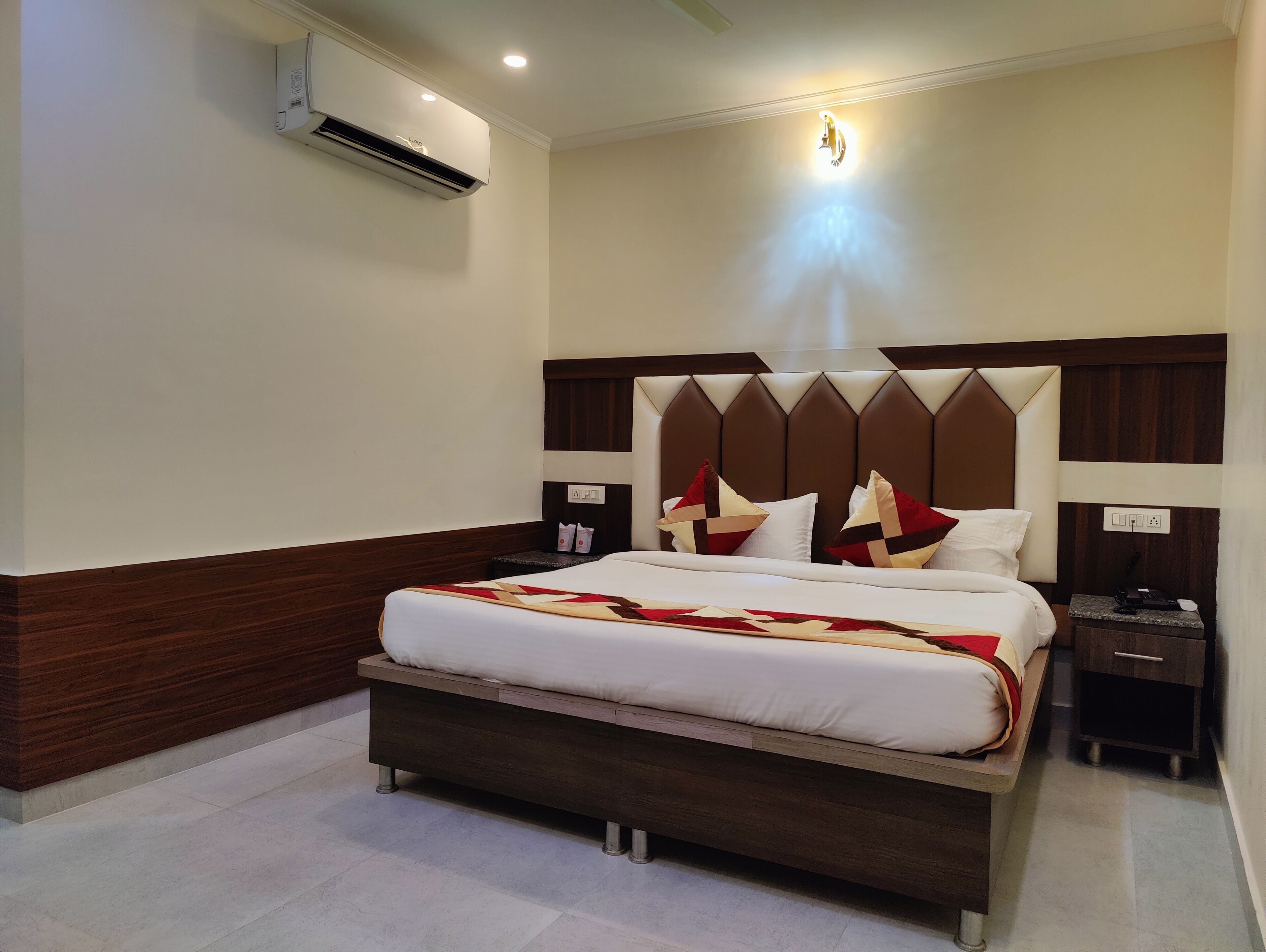 A hotel room with a bed, bedside tables, an air conditioning unit, and a wall-mounted light fixture.