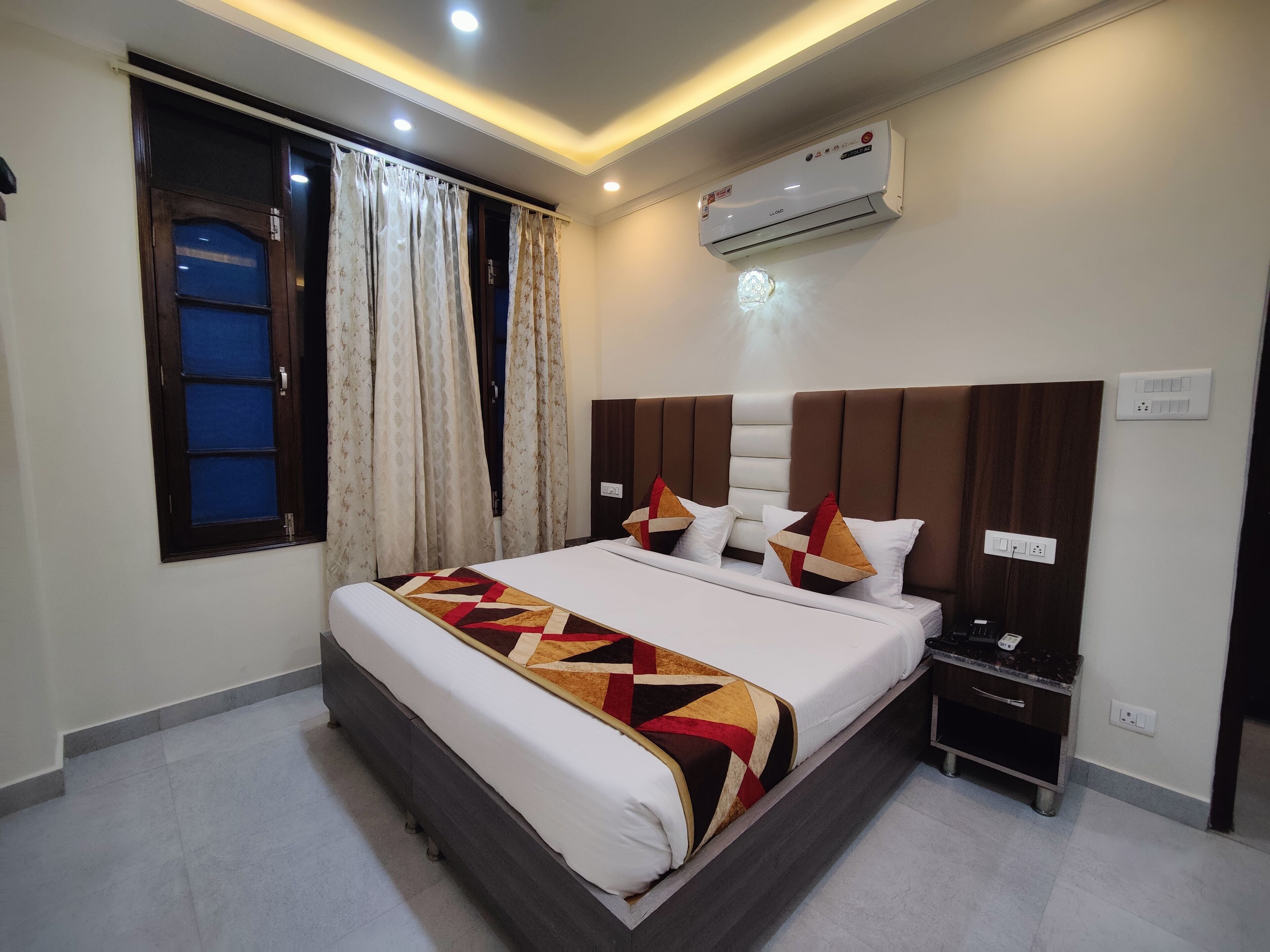 A hotel room with a large bed, an air conditioner, a bedside table, and a window with curtains.