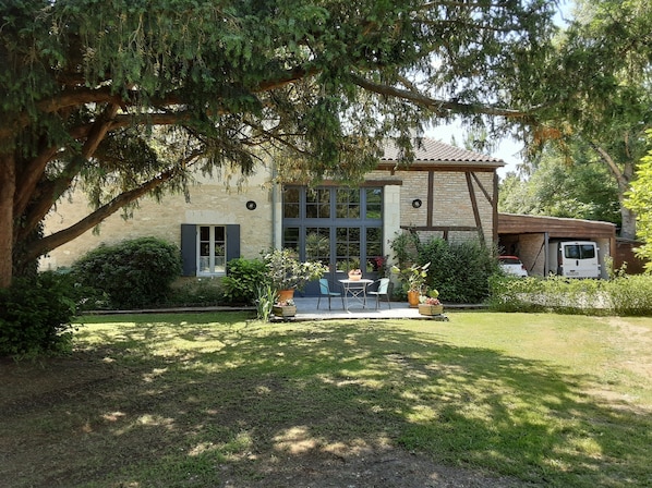 Une maison avec une façade en pierre, un grand arbre dans le jardin et une terrasse avec des meubles d’extérieur.
