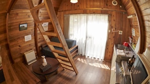 Interior de una cabaña de madera con una cama en altillo, una mesa de comedor, una zona de cocina y una ventana con cortinas.