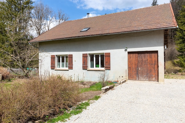 A single-story house with a sloped roof, two windows with shutters, a wooden door, and a gravel driveway.