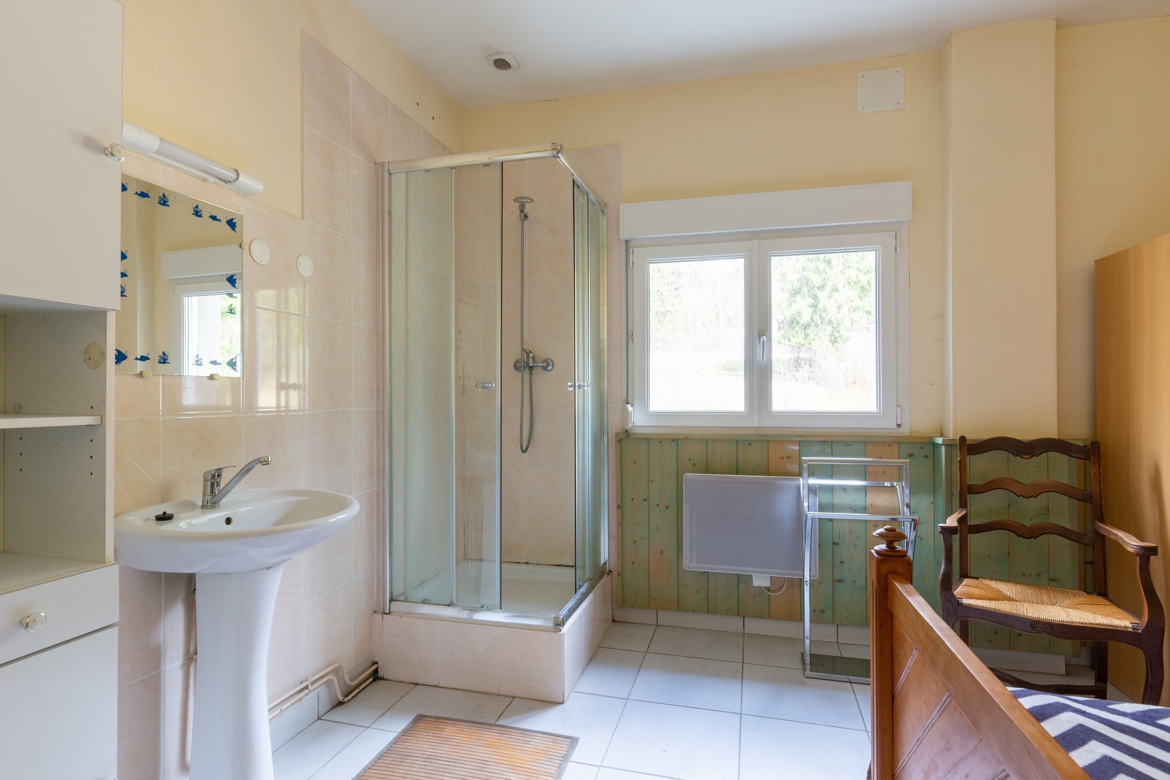 Ein Badezimmer mit einer Duschkabine, einem Waschbecken, einem Holzstuhl und einem Fenster.
