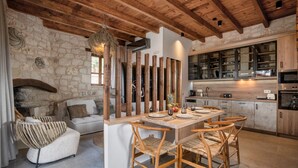 Un interior rústic amb paret de pedra, sostre de fusta, zona de menjador amb taula i cadires de fusta, sofà, i cuina amb armaris i electrodomèstics.