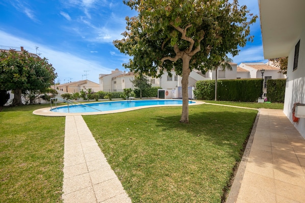 Piscina com calçada pavimentada, rodeada por grama bem cuidada e uma árvore.