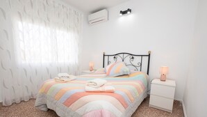 Un lit bien fait, recouvert d’une couverture à rayures, une table de chevet avec une lampe et un climatiseur fixé au mur.