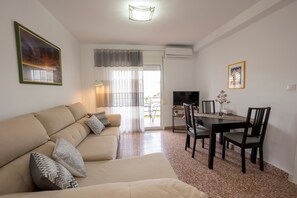 Sala de estar com sofá, mesa de jantar e cadeiras. Há uma televisão e uma janela com cortinas.