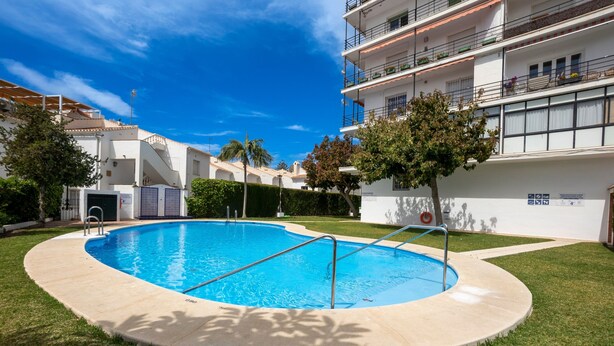 Une piscine avec un escalier en métal, entourée d’une pelouse et bordée d’un bâtiment blanc doté de balcons.