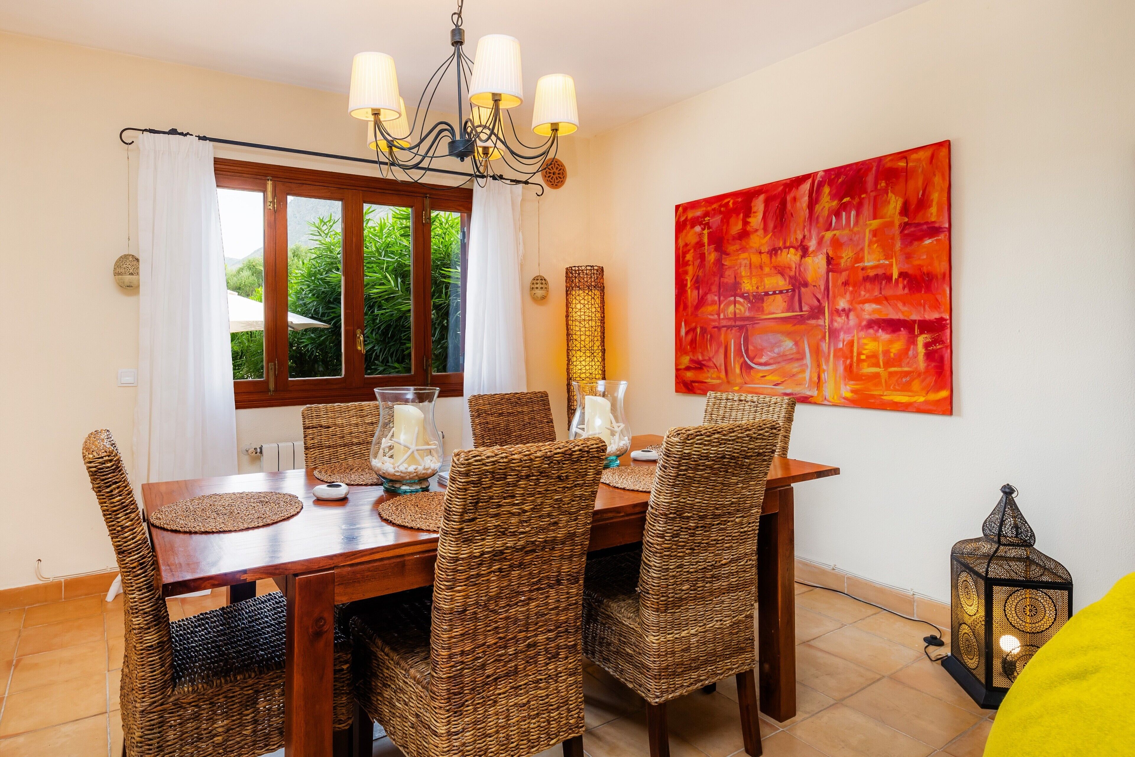 Un coin repas avec une table en bois, des chaises en osier, un lustre et un grand tableau abstrait.