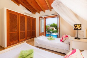 A room with two beds, a window with a view of a pool and mountains, wooden furniture, and a ceiling fan.