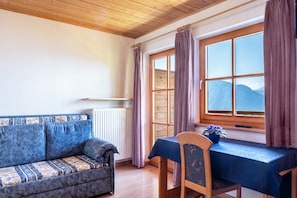 Una stanza con soffitto in legno, un divano, un tavolo con una sedia, una finestra con tende e un termosifone.