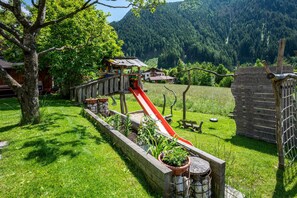 A wooden play structure with a slide, surrounded by a grassy area with trees and a mountain in the background. - Studio Apartment "Talblick 2" with Mountain View, Garden and Wi-Fi (Lajen)