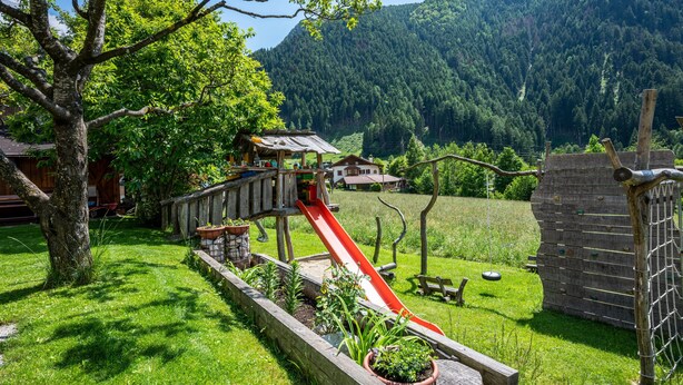 Eine hölzerne Spielanlage mit Rutsche, umgeben von einer grünen Wiese mit Bäumen und einem Berg im Hintergrund.
