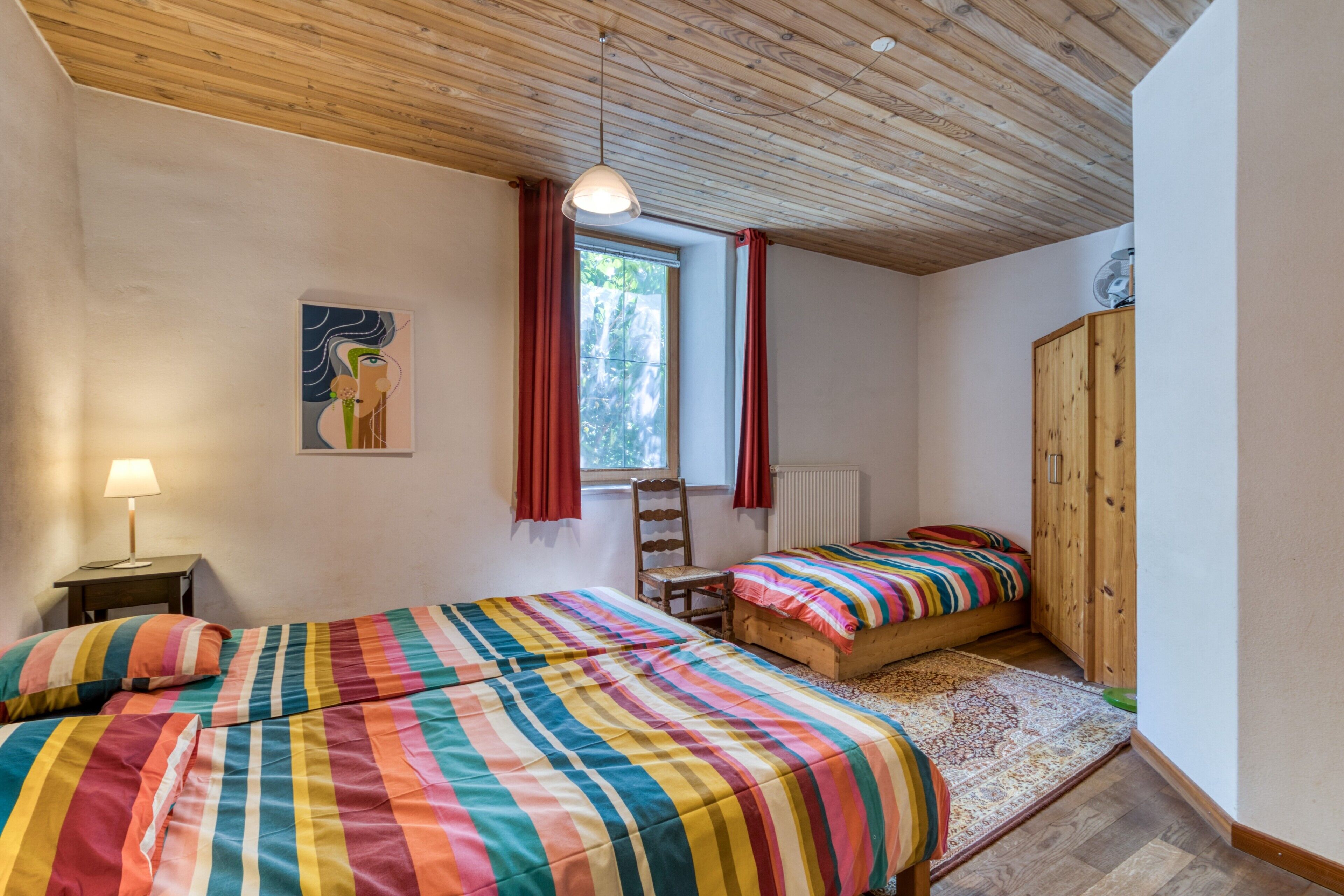 A bedroom with two beds, a wooden ceiling, a window with red curtains, a chair, and a cabinet.