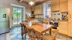 Een keuken met houten kasten, een eettafel, stoelen, een koelkast, een magnetron en een klok aan de muur.