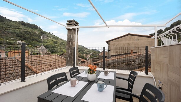 A terrace with a table and chairs, a view of a hilly landscape, and a building in the background.