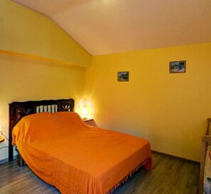 Ein Schlafzimmer mit einem Bett, das mit einer orangefarbenen Decke bezogen ist, einem Nachttisch mit einer Lampe sowie zwei kleinen Bildern an der Wand.
