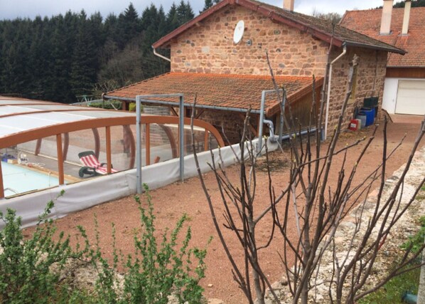 A stone building with a tiled roof, a swimming pool with a glass enclosure, and a garden area with bare branches.