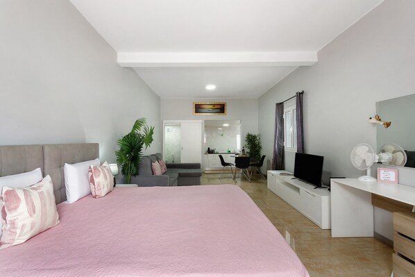A modern bedroom with a bed, a desk, a television, and a dining area in the background.