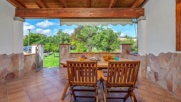 An outdoor patio with a wooden table and chairs, stone walls, and a view of greenery and buildings.