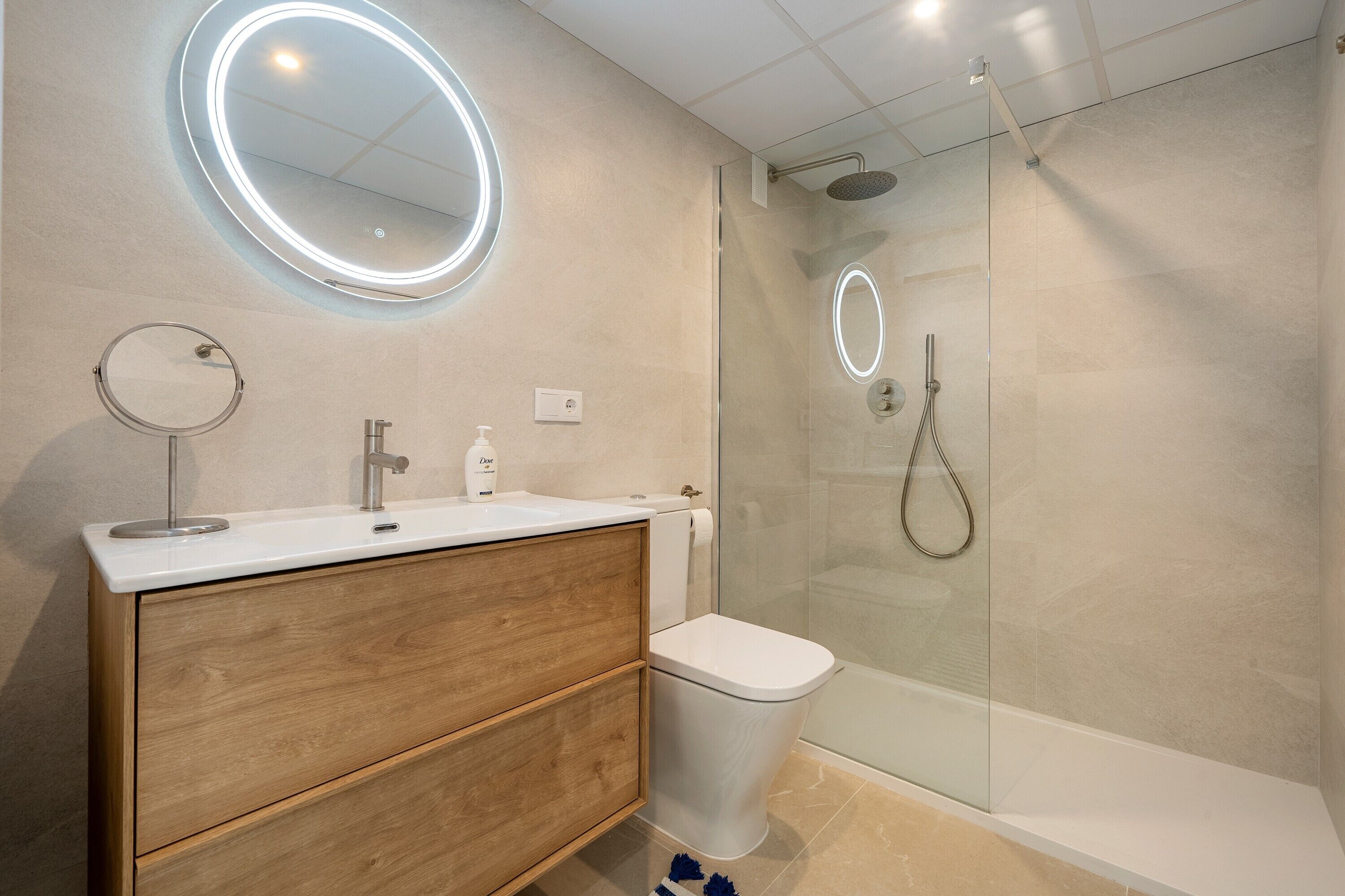 Een moderne badkamer met een houten wastafel, een ronde spiegel, een toilet en een douchecabine met glazen wanden.