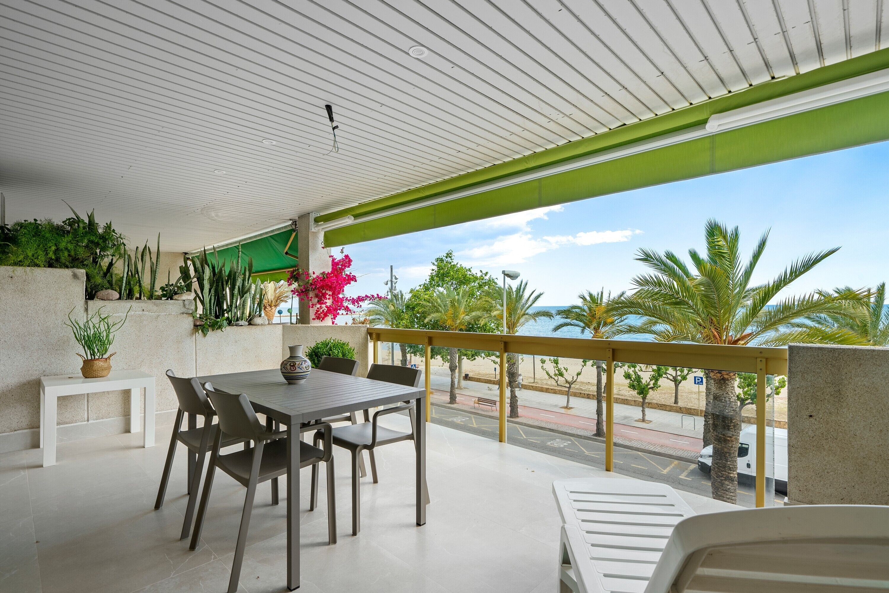 Un espacio comedor al aire libre con techo, una mesa y sillas, plantas en macetas y vista a la playa y palmeras.