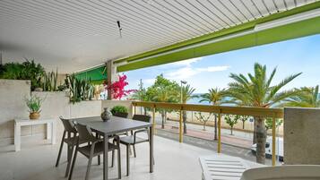 Un espacio comedor al aire libre con techo, una mesa y sillas, plantas en macetas y vista a la playa y palmeras.
