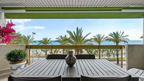A covered outdoor seating area with a table and chairs, a decorative vase, and a view of the beach and palm trees. - Apartment 'Pelicano' with Sea View, Wi-Fi and Air Conditioning (Cambrils)
