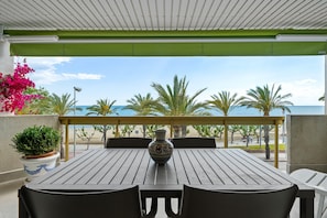 A covered outdoor seating area with a table and chairs, a decorative vase, and a view of the beach and palm trees.
