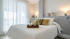 A modern bedroom with a large bed, a bedside table with lamps, and a view of the outdoors through sheer curtains. - Apartment 'Pelicano' with Sea View, Wi-Fi and Air Conditioning (Cambrils)