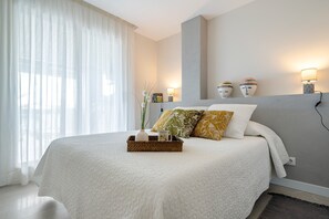 Un dormitorio moderno con una cama grande, una mesita de noche con lámparas y vistas al exterior a través de cortinas translúcidas.