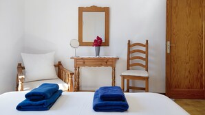 Un dormitorio con una cama, dos toallas azules, una silla de madera, una mesita de noche con un jarrón, un espejo y una puerta de madera.