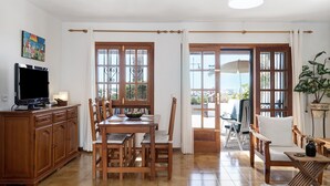 Un comedor con mesa y sillas de madera, un televisor sobre un mueble bajo, y ventanas amplias con cortinas.